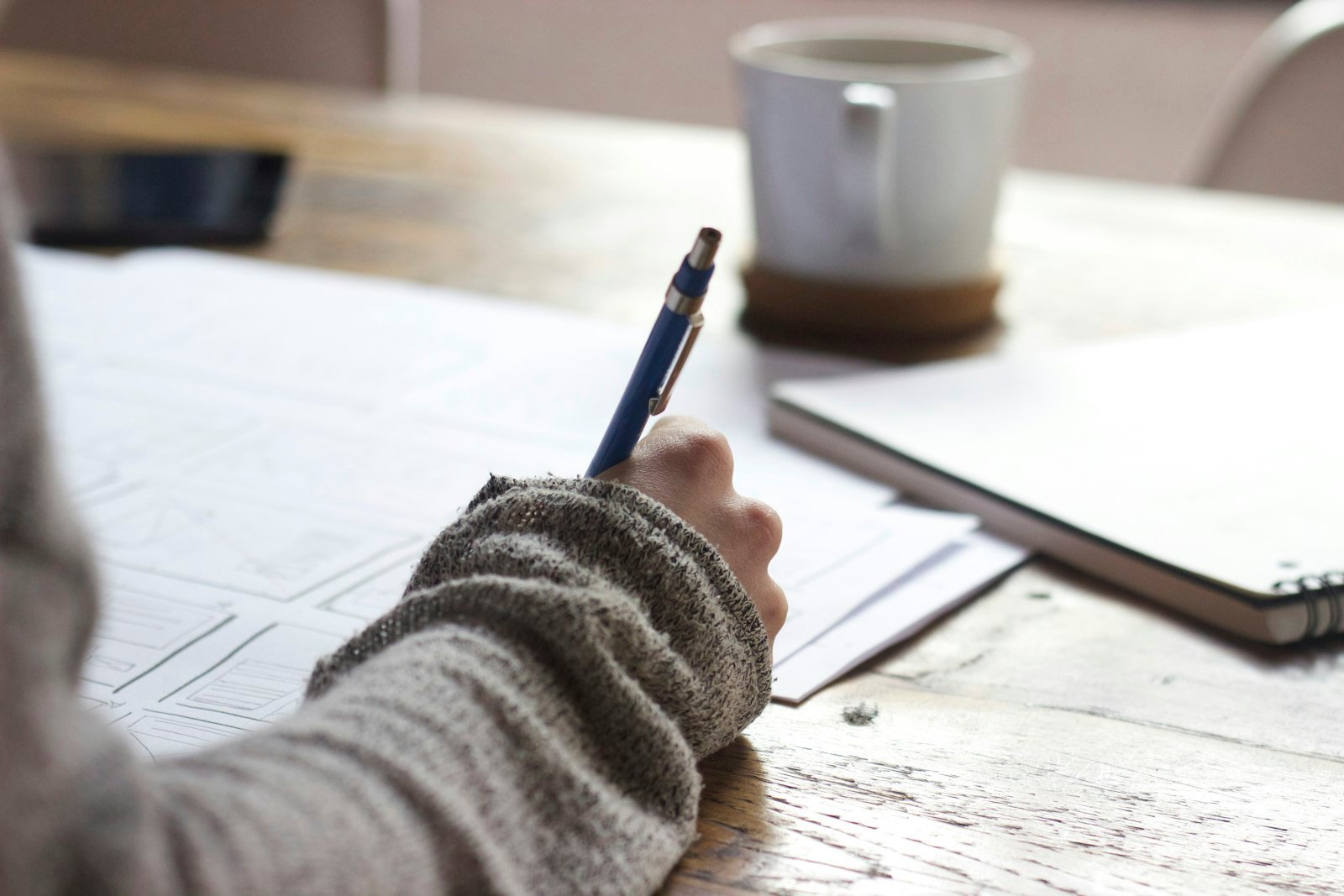 Dłoń z niebieskim długopisem nad kartką, w tle kubek i notes